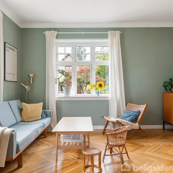 En stue med et grønt vægtema. En lyseblå sofa er placeret ved siden af et vindue med planter. En trækommode og stol kompletterer rummet. Solsikker står i vindueskarmen.