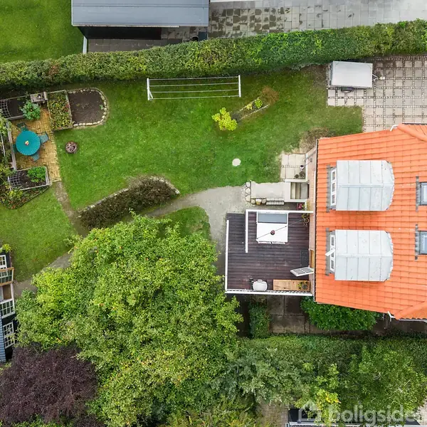 Hus med rødt tag og to ovenlysvinduer, set oppefra. Haven har en terrasse, græsplæne og en udendørs spiseplads med parasol. Stier og hække omkranser området.
