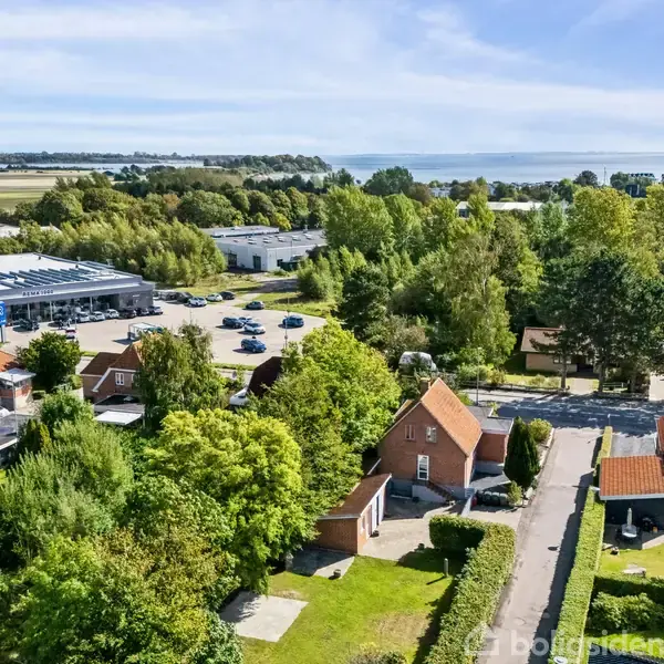 Huse med røde tage står blandt grønne træer; en parkeringsplads ligger bagved. I baggrunden ses åbne marker og havudsigt under en blå himmel.