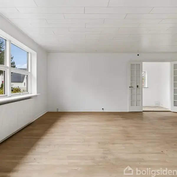 Lyst, tomt rum med hvidmalede vægge og trægulv. Store vinduer til venstre giver udsigt til et hus og træer udenfor. En åben dør med glasruder fører til et andet rum.