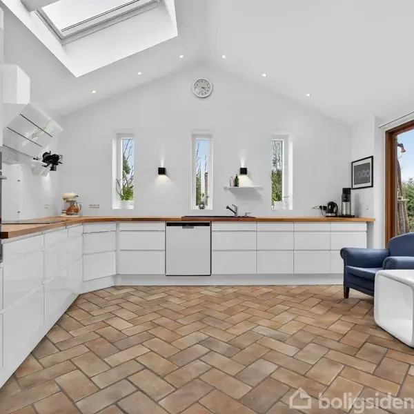 Et moderne køkken domineret af hvide skabe og en træbordplade, med indbyggede ovne. Der er en skrånende loft med ovenlys. Ved siden af står en blå lænestol og hvid puf.