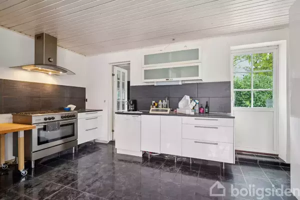 Køkken med hvidt kabinet og moderne komfur under en emhætte. Glasdør til højre og et vindue med udsigt til grønne træer. Sorte fliser på gulv og væg.