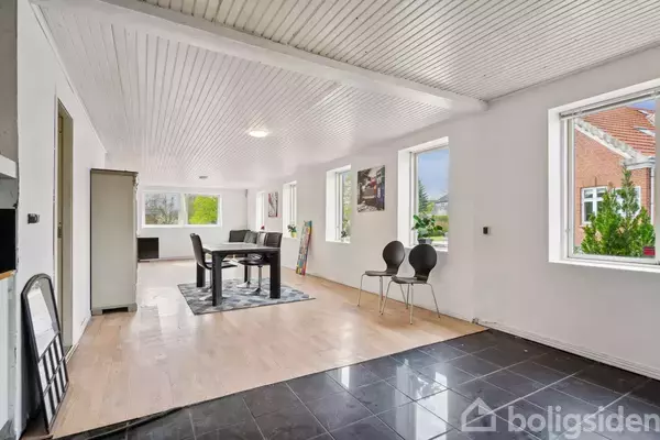 Spisebord med stole står på et tæppe i en lys stue med lyst træ- og sort flisegulv, hvide vægge og vinduer mod en have med buske og træer.