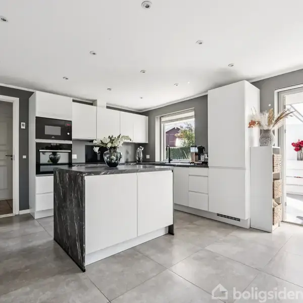 Et moderne køkken med en mørk marmorbordplade på en central ø. Køkkenet har hvide skabe, integrerede apparater og store vinduer. En dør åbner ud til en lys terrasse.