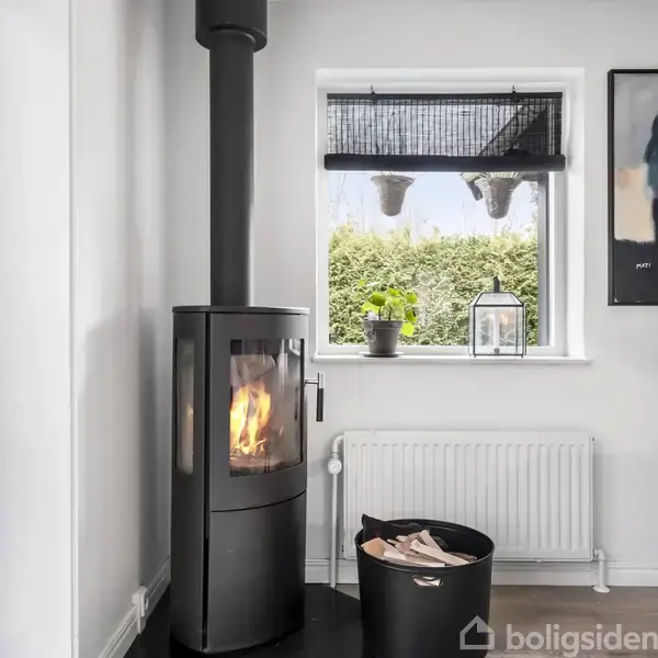 Brændeovn med levende ild står ved siden af et vindue. Udenfor ses grønne buske. En plante og en lanterne er på vindueskarmen. En radiator er placeret under vinduet.