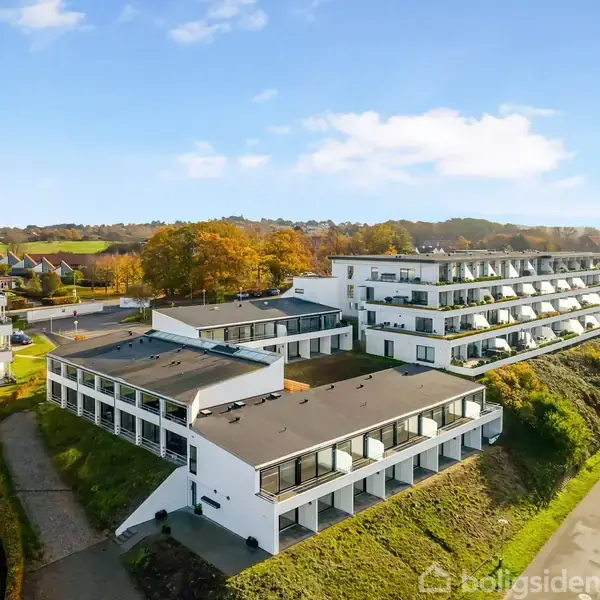 Et moderne hvidt lejlighedskompleks med flere etager ligger på en grøn bakkeskråning. Det har store balkoner, og i baggrunden ses flere træer og et boligområde.