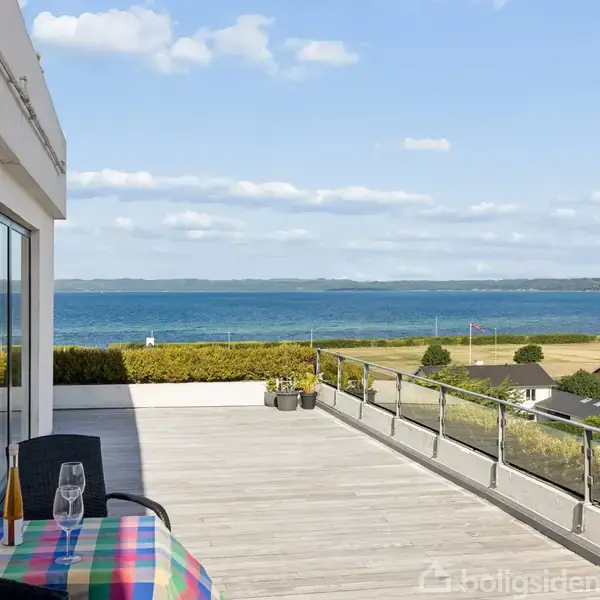 En terrasse med glasskærm udstyret med et bord med farverigt tæppe og stole. Et vinflaske og glas står på bordet. Udsigt over havet og landskab i baggrunden.