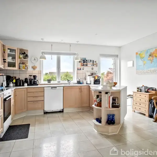Køkken med træskabe og hvidevarer arrangeret langs væggene. Et rundt bord står midt i rummet. Vinduer giver udsigt til træer. Kort på væggen og en træstol til højre.
