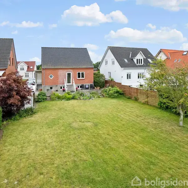 Rødt murstenshus med have og terrasse ligger i et kvarter med lignende huse. Flere huse ses i baggrunden, omgivet af grønne haver og klare himmel.