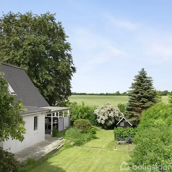 Et hus med skråtag står stille i en have omgivet af træer og buske. I baggrunden ses en åben mark under en klar, blå himmel.