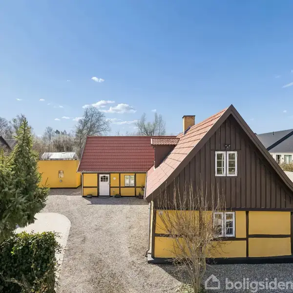 Et gult bindingsværkshus med rødt tegltag står på en grusbelagt grund. Omkring huset ses andre huse, og træer markerer en del af ejendommens grænse.