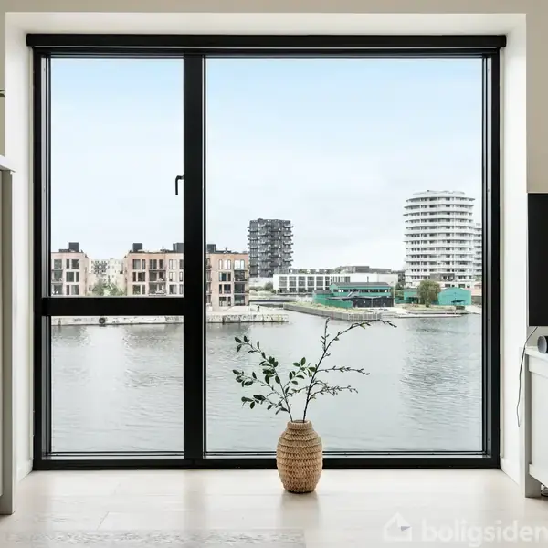 Et stuevindue viser en udsigt over vandet med moderne lejlighedsbygninger i baggrunden. Foran vinduet står en plante i en kurv. Møbler omfatter et hvidt skab og TV.