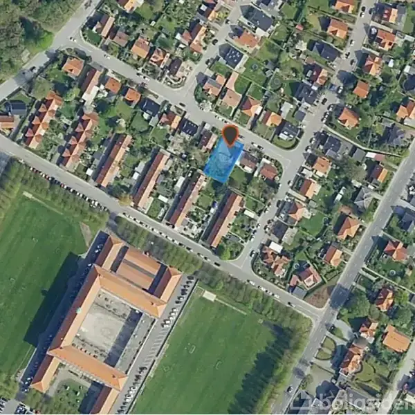 Luftfoto af boligområde, hvor en orange markør peger mod et specifikt hus omgivet af flere rækkehuse og grønne områder. Veje og et større bygningskompleks ses i nærheden.