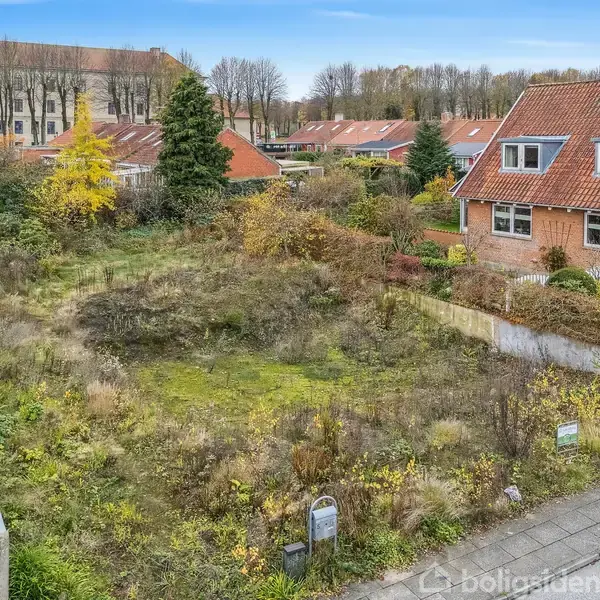 En tom grund dækket af vildtvoksende buske og græs ligger mellem to huse i et villakvarter. I baggrunden ses flere hustage og et større institutionsbyggeri.