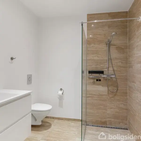 Badeværelse med bruser bag glasvæg til højre, hvidt toilet og håndvask til venstre. Væggene er hvide og brusenichen har brune fliser. Enkelt og moderne design.