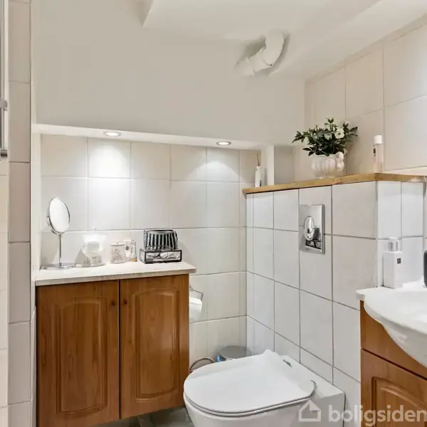 Badeværelse med toilet og vask, der er placeret op ad flisevægge. En bruser med rødt brusegel ses til venstre. Et indbygget skab med trædøre indeholder toiletartikler.