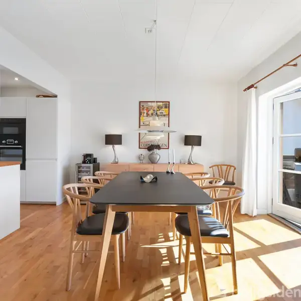 Et moderne spisebord omgivet af seks stole står på et trægulv. Baggrunden har en lys væg med kunst, to sorte lamper og franske døre, der lader sollys ind i rummet.