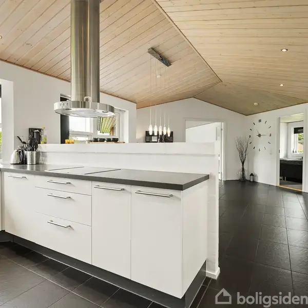 Hvidt køkken med moderne design, skarpt afgrænset af en kogeø, står i lyse omgivelser med sort gulv og træloft. Store vinduer lukker naturligt lys ind i rummet.