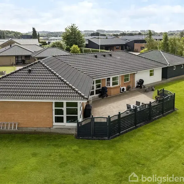 Et murstenshus med mørkt tag står centralt. Det omkranser en flisebelagt terrasse med havemøbler. Huset er placeret i et velplejet, grønt boligkvarter med naboejendomme synlige i baggrunden.