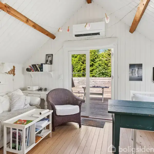 En lys stue med sofa, stol og bord åbner mod en terrasse gennem glasdøre. Hvide vægge og loft med træbjælker giver en hyggelig atmosfære. Billeder og bøger dekorerer rummet.