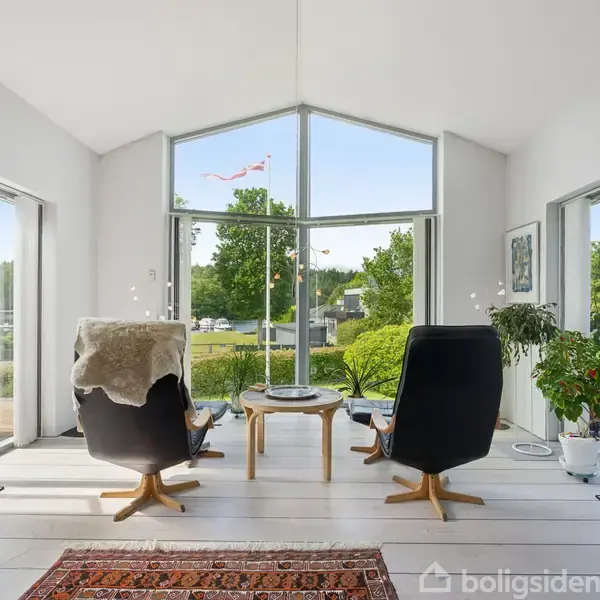 To komfortable sorte lænestole vender mod et stort vindue med udsigt til en have. Ved siden af står en grøn plante, og en farverig tæppe pryder det lyse trægulv.
