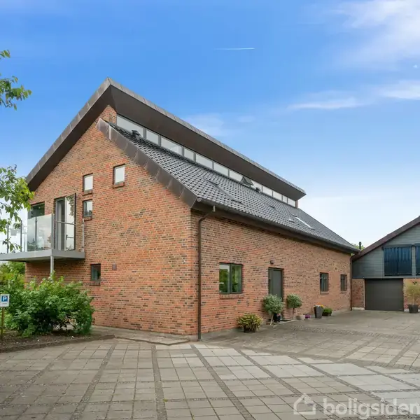 Et moderne murstenshus med skråt tag og en balkon til venstre. Det står på en flisebelagt gårdsplads omgivet af grønne buske og træer under en klar blå himmel.