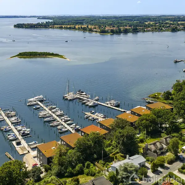 Havneområde med fortøjede både langs flere moler. Omgivelserne har tæt bevoksning og bygninger med røde tage. Længere ude ses en lille, grøn ø midt i vandet.