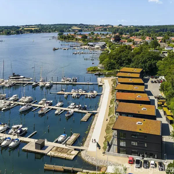 Både ligger i en marina ved vandet, med moderne bygninger langs kajen. Landsby og skov klæder baggrunden under en blå himmel.