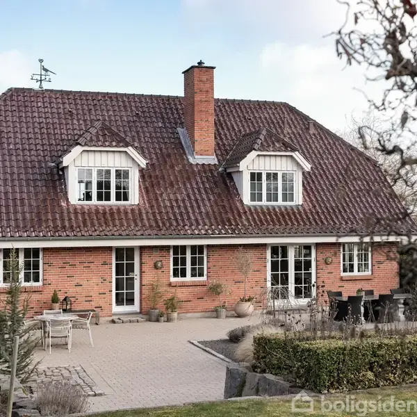 Rødt murstenshus med brun teglstenstag, to kviste, og en centralt placeret skorsten. Foran huset er en flisebelagt terrasse med havemøbler. Omgivelserne inkluderer træer og en velplejet have.