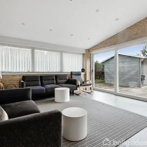 En stue med sort sofa står stille i et lyst rum med store vinduer, der giver udsigt til en terrasse og en garagebygning. Et vægur hænger på væggen.