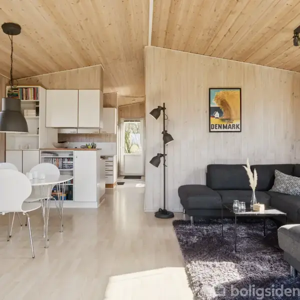 Sort sofa står i stue med trævægge og -loft ved siden af et lille sofabord med vaser. Spisebord med hvide stole står tæt ved. Væggene har en plakat med teksten "DENMARK."
