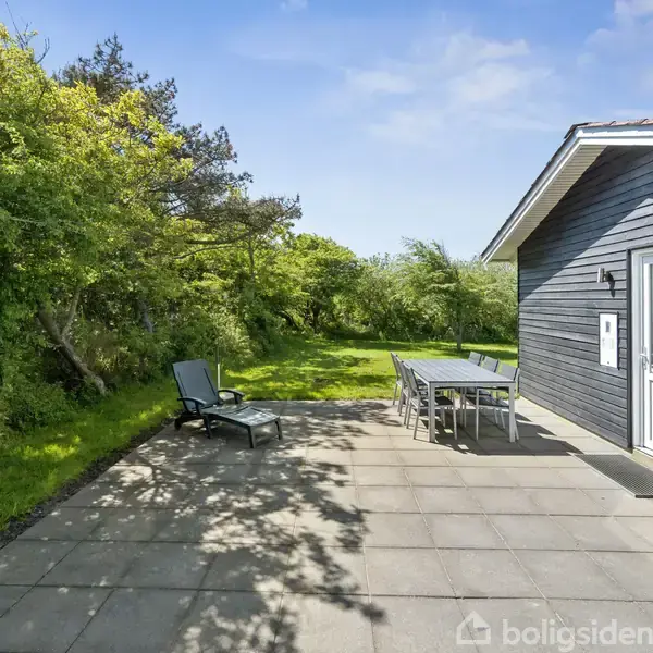 En mørkeblå træbygning med en dør vender ud mod en flisebelagt terrasse. En sort liggestol og et spisebordssæt står under åben himmel omgivet af grønne buske og træer.