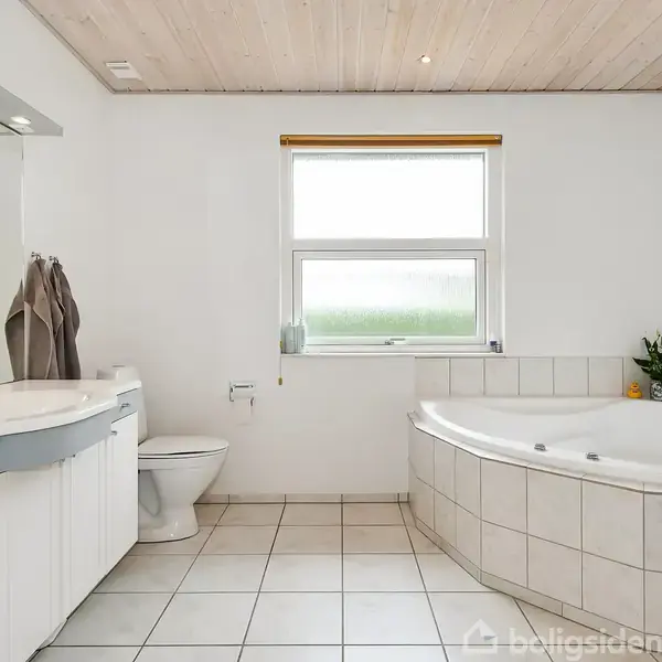 Et rummeligt badeværelse med stort hjørnebadekar og vask ved siden af et toilet. Lyst gulvfliser, hvid væg og et vindue med rullegardin skaber en luftig atmosfære.