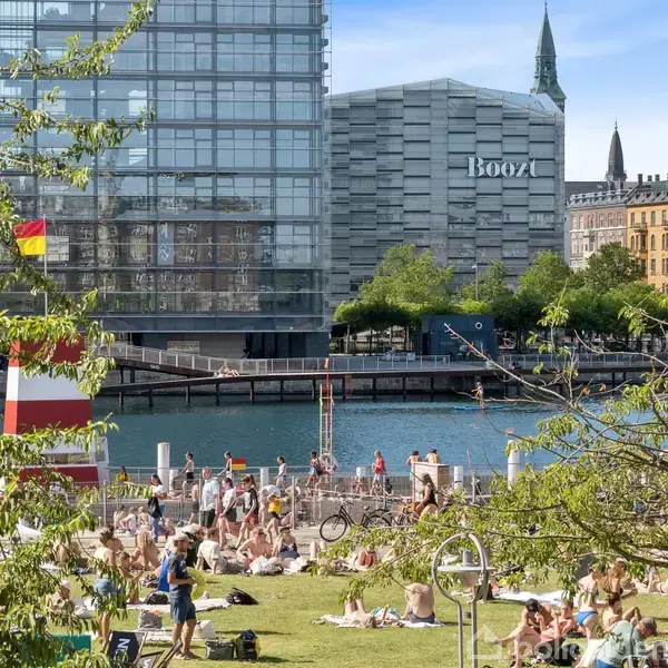 Mennesker soler sig og slapper af på en græsplæne ved en vandkant. Baggrunden viser moderne bygninger, heriblandt en med teksten "Boozt", og et tårn ses i horisonten.