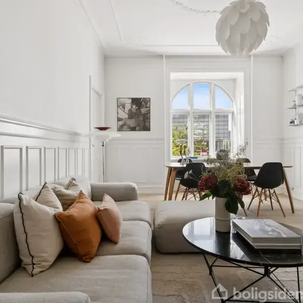En lys stue med en beige sofa, et rundt sofabord med bøger og blomster, og spisebord med stole i baggrunden. Store vinduer giver udsigt til grønne træer udenfor.