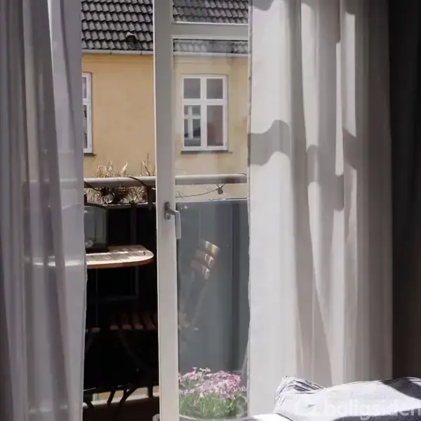 Glasdør står åben til en altan med et lille bord ved siden af en gelænder. Udsigten viser en beige bygning med to vinduer.