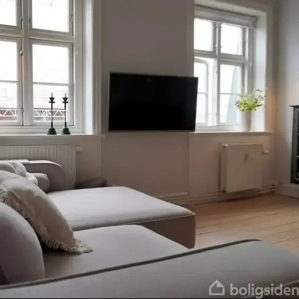 En grå sofa står i en lys stue ved vinduer, mens en fladskærm hænger på væggen. En skænk med glaslåger ses til højre. Ingen tekst er synlig.