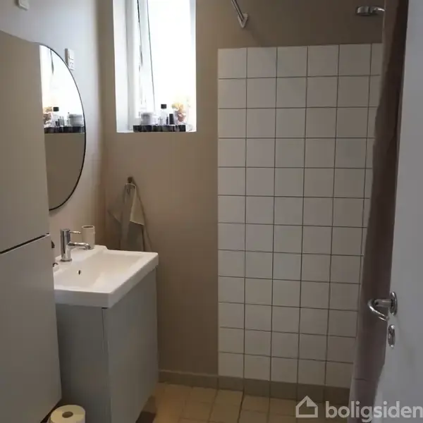 En håndvask med spejl hænger på en lysebrun væg. Ved siden af er hvide fliser. Ovenpå vasken står forskellige flasker ved et lille vindue. En håndklædeholder er ved siden af.