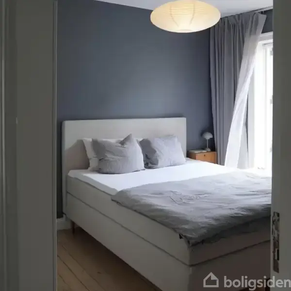 En seng står i et soveværelse med blå vægge og grå gardiner. Der er puder på sengen og et natbord med lampe ved siden.