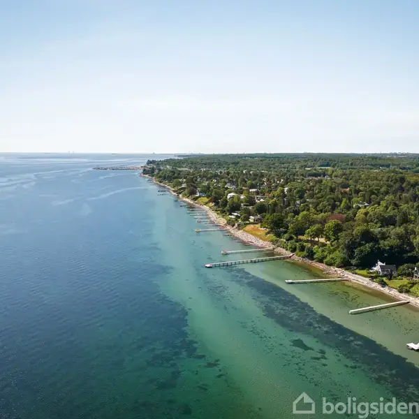 Kystlinje ved havet under en klar himmel. Langs kysten løber en række bådebroer, omgivet af tæt bevoksning. Husområder kan skimtes i baggrunden.