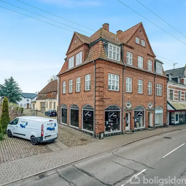 Rødt murstensbygning med skråtag står på en gadehjørne. Biler er parkeret på asfalten ved siden af bygningen, omgivet af andre bygninger og en blå himmel.