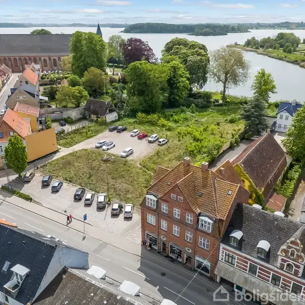 En åben parkeringsplads med flere biler står blandt bygninger i en by. En stor kirke ses i baggrunden, omgivet af træer nær en sø.
