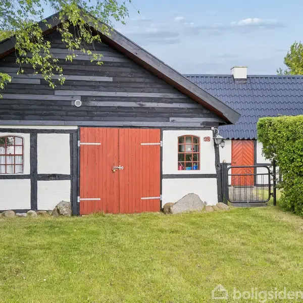 En bindingsværksbygning med hvide og sorte vægge, og røde døre står stille omgivet af grønne buske og en velholdt græsplæne under en klar himmel.