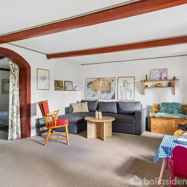 Stue med sofa og sofabord ved siden af en rød gyngestol. Gardiner adskiller stuen fra et tilstødende soveværelse. Rummets vægge er prydet med kunstværker, og en lille hylde hænger over sofaen.