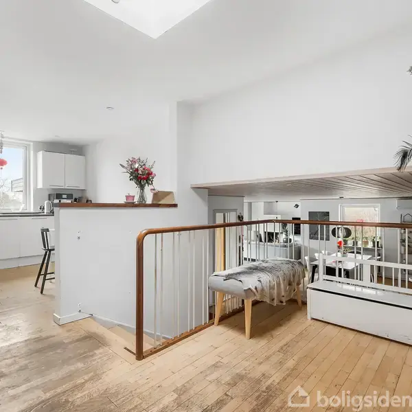 Køkken med hvide skabe og store vinduer, som giver lys til rummet. Trægulve forbinder køkkenet med et lavere stueområde med trægelænder og dekoration med planter og blomster.