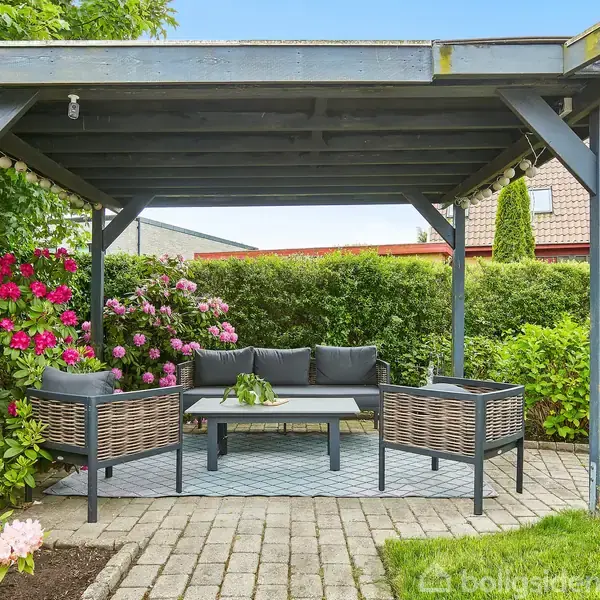 En udendørs siddegruppe med to stole og en sofa står under en pergola. Der er brosten på jorden, omgivet af blomstrende buske og en velplejet have.