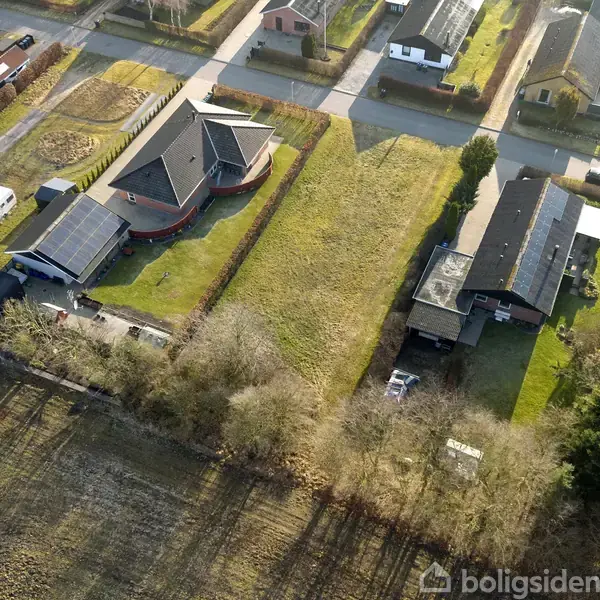 To huse med solpaneler på tagene står i en forstad med velholdte græsplæner. Omgivet af flere huse langs en asfaltvej med træer og buske omkring.