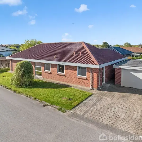 Rødt murstenshus med mørkt tegltag står ved siden af en garage. Beliggende på en stille villavej med græsplæne foran og et enkelt træ til venstre.