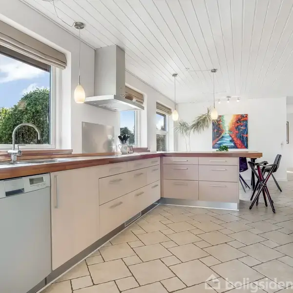 Et moderne køkken præsenterer indbyggede hvidevarer og lyse trælister, med to store vinduer, der slipper naturligt lys ind. I baggrunden er et spiseområde med farverig kunst på væggen.