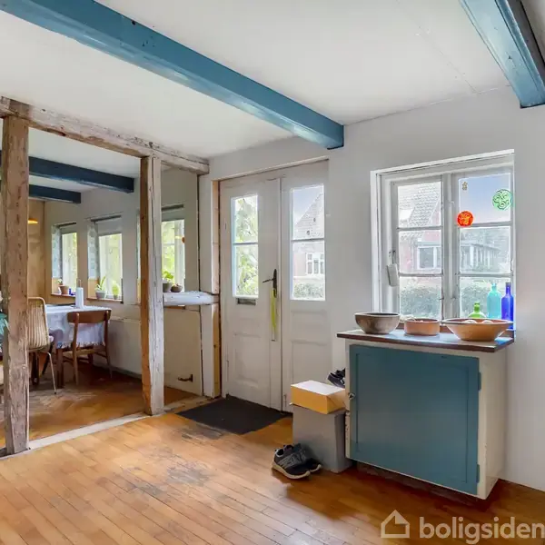 Rum med trægulv og loftsbjælker, flere vinduer og dør mod en terrasse. Et spisebord med stole står ved siden af en plante. En kommode med skåle står under et vindue.